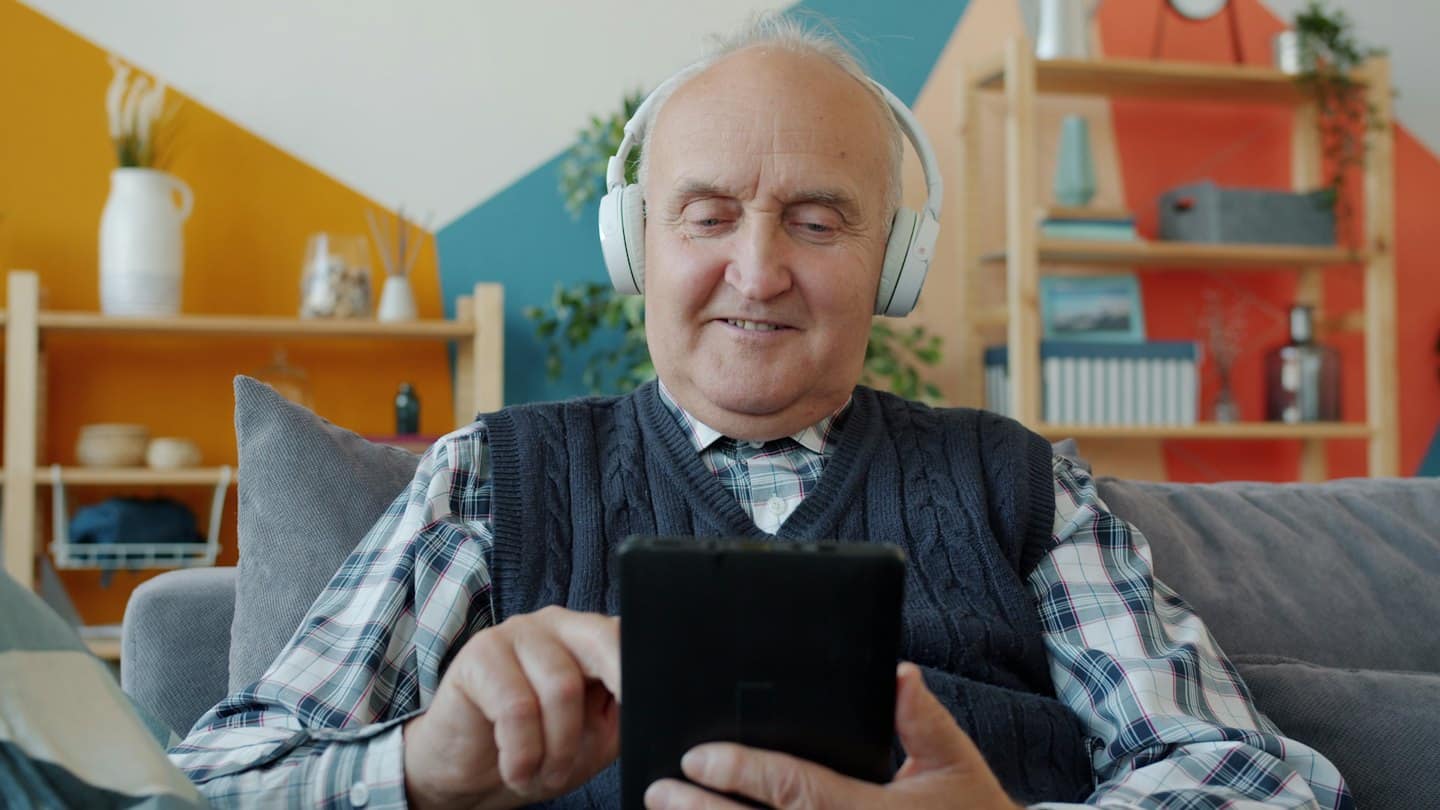 Äldre man med hörlurar ler när han använder surfplatta i soffan.