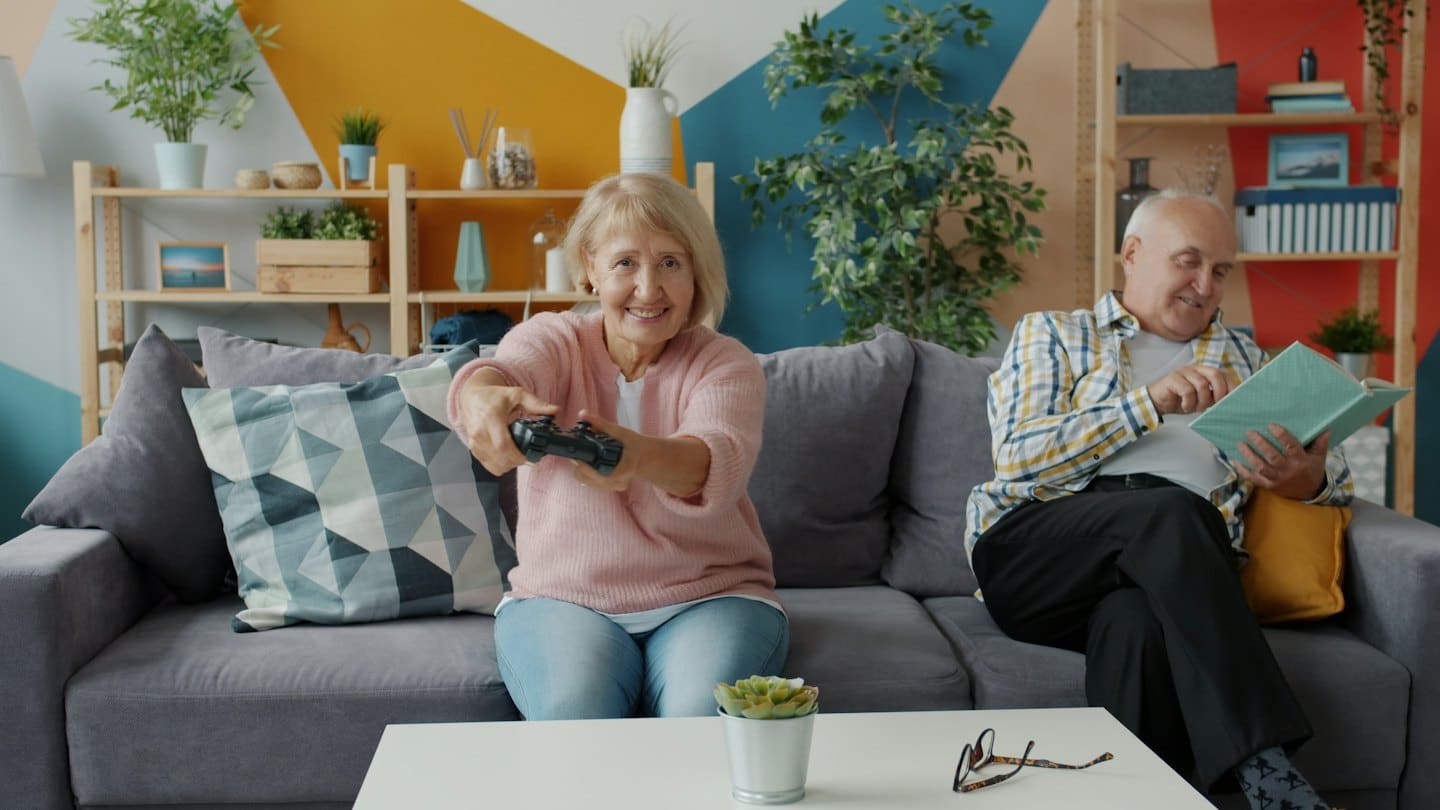 Äldre kvinna spelar tv-spel i soffan medan en man läser.